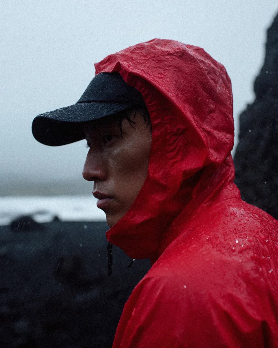 A man in a red rain jacket and black cap on a wet, grey shoreline.
